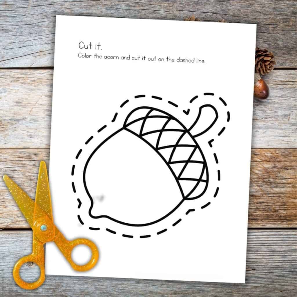 Printable preschool worksheet featuring an acorn to color and cut out along the dashed lines, with scissors on a wooden surface.