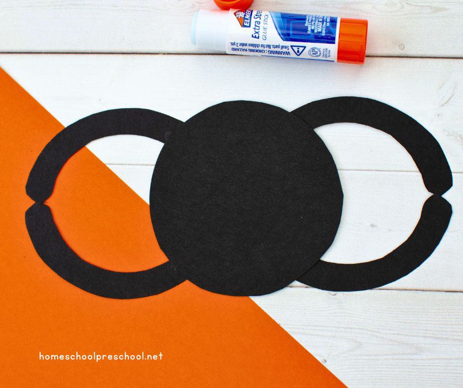A partially assembled paper spider with glued legs attached to a round black body, with a glue stick nearby on a white and orange surface.