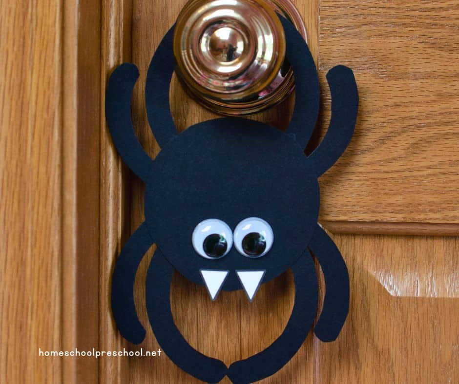 A completed paper spider craft with googly eyes and white fangs hangs on a door handle, creating a fun Halloween decoration.
